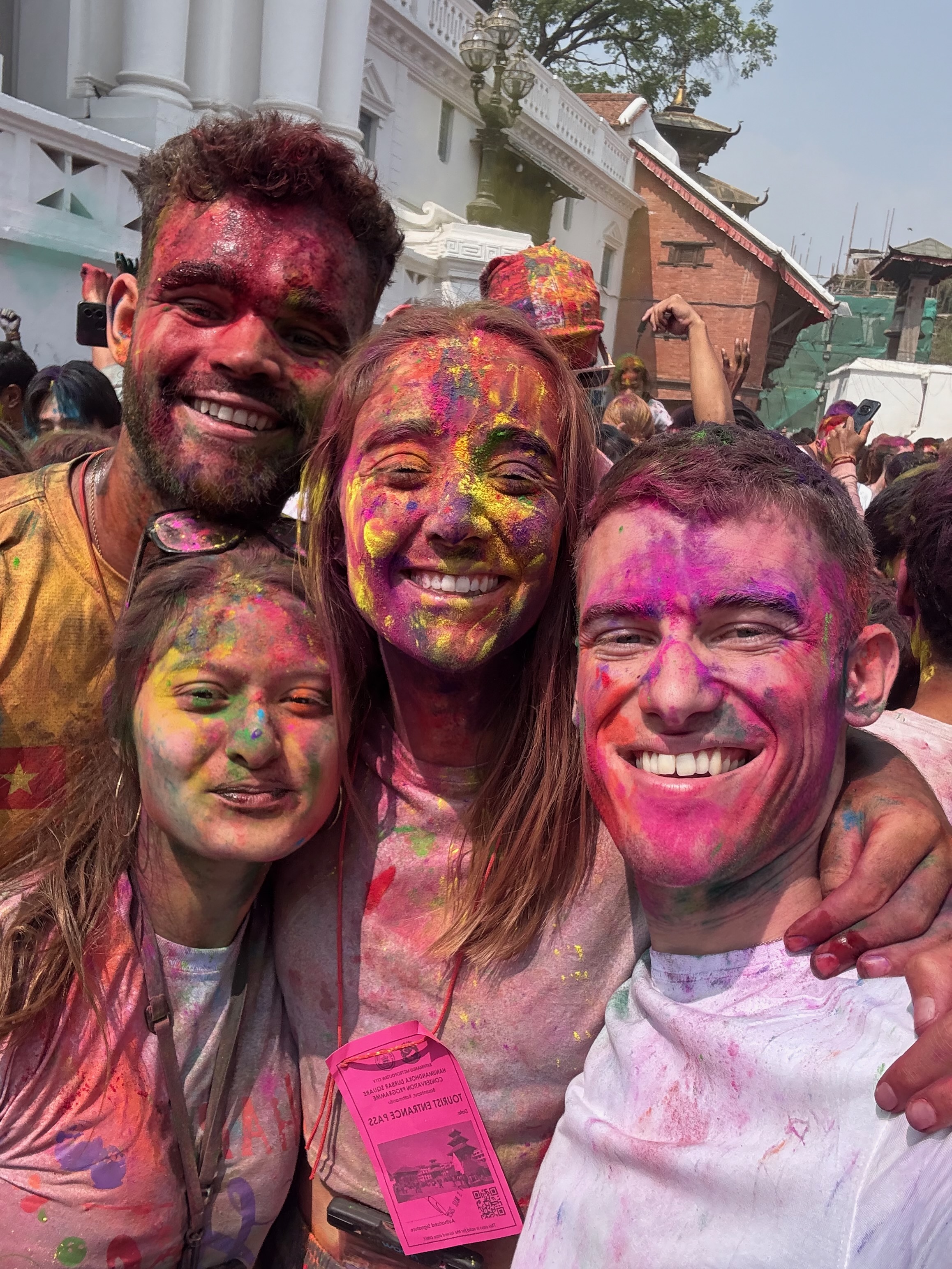 A group of friends laughing and covered in vibrant pink, blue, and yellow Holi powders while standing outdoors in Nepal.