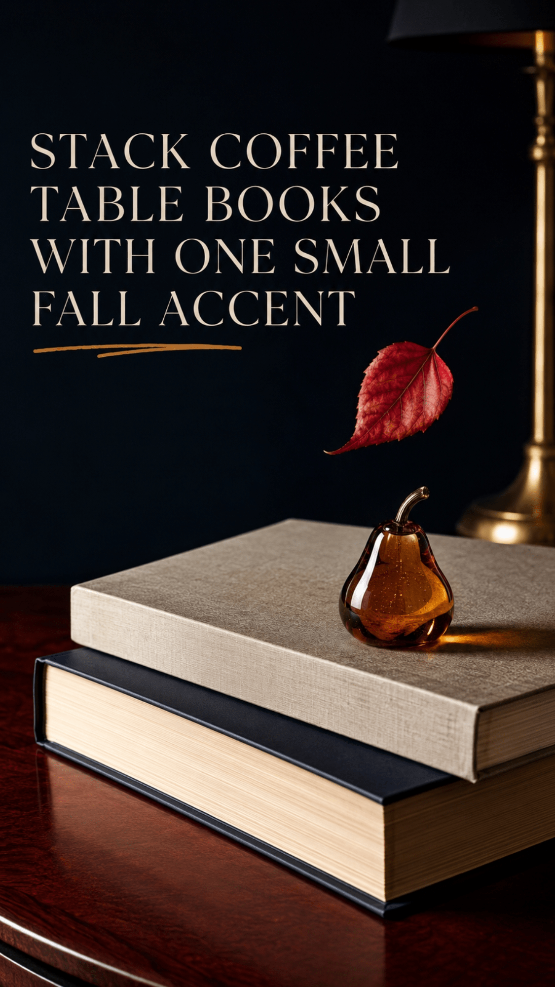 Stacked coffee table books topped with a small amber glass pear beside a brass lamp and floating red autumn leaf.