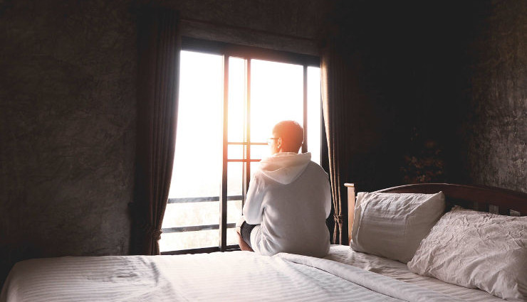 Back view of young man with eyeglasses sitting alone on bed and looking through the window in bedroom, vintage filter effect.