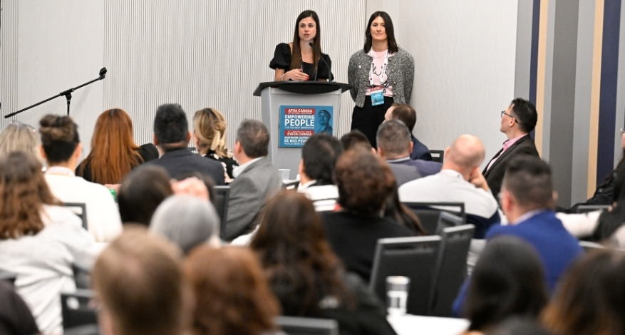 Two women stand at a podium presenting in front a seated crowd.