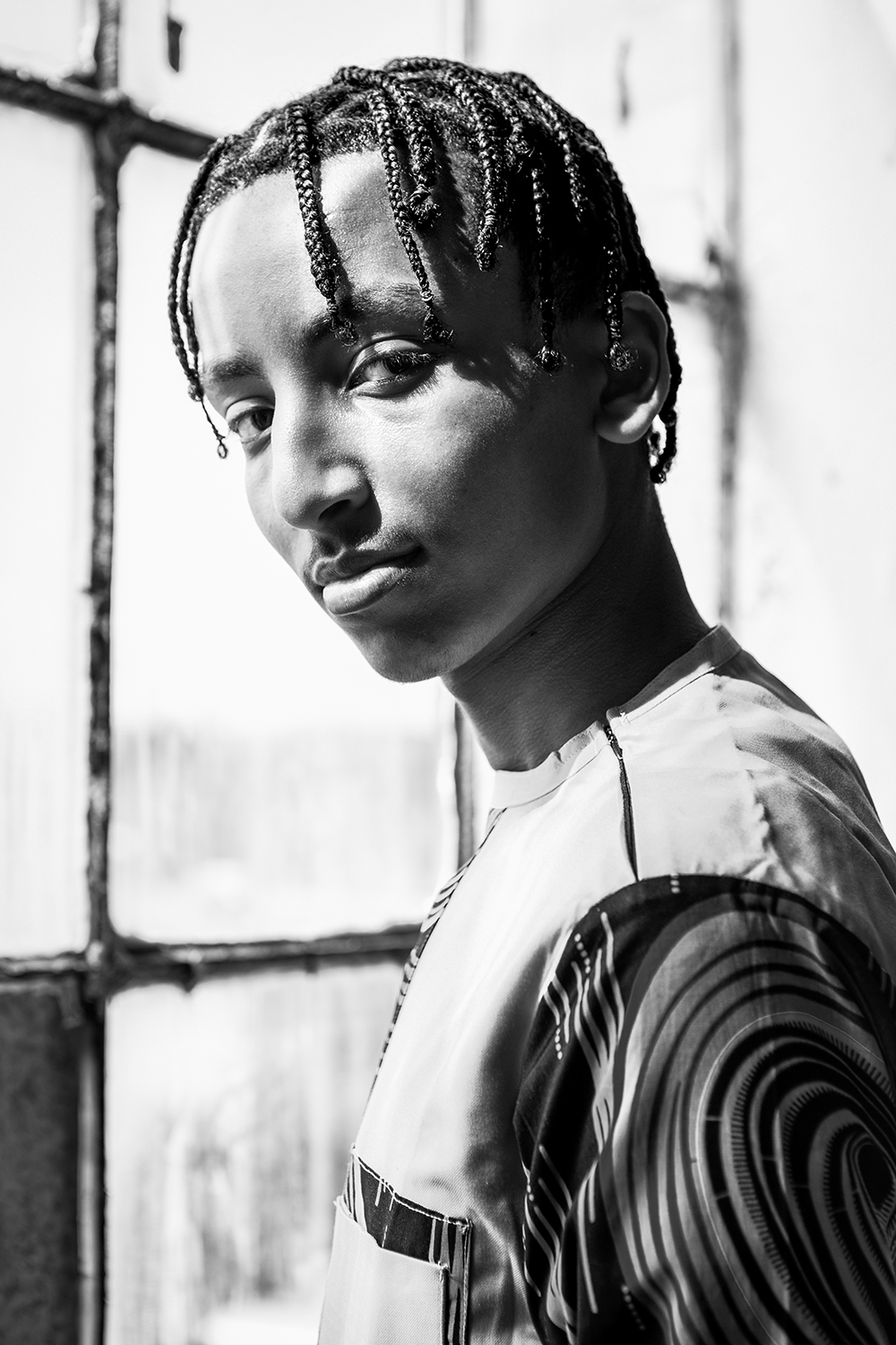 Afrikanischer junger Mann mit Braids Frisur und traditioneller Kleidung, steht vor einem Industriefenster. Das Bild ist schwarz-weiß. Er blickt seitlich Zur Kamera