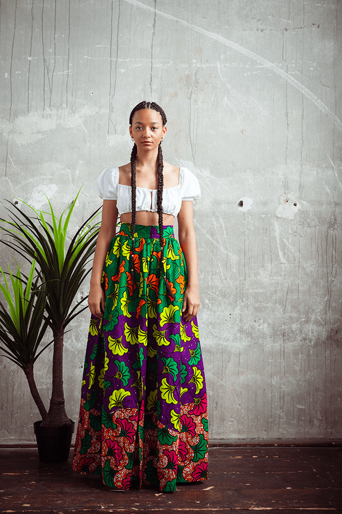 afrikanische Frau mit langen geflochtenen Zöpfen, Sie trägt ein traditionelles afrikanisches Gewand, in einem Industriegebäude. Sie steht vor einer Betonwand neben einer Pflanze und blickt gerade in die Kamera