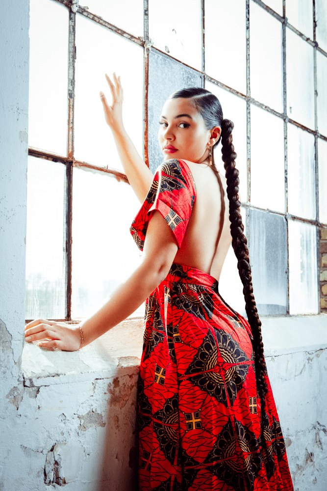 Afrikanische Frau steht am Fenster, die Haare sind zu einem dicken Zopf geflochtene trägt ein traditionelles, afrikanisches rotes Kleid und trägt große Kreolen Ohrringe