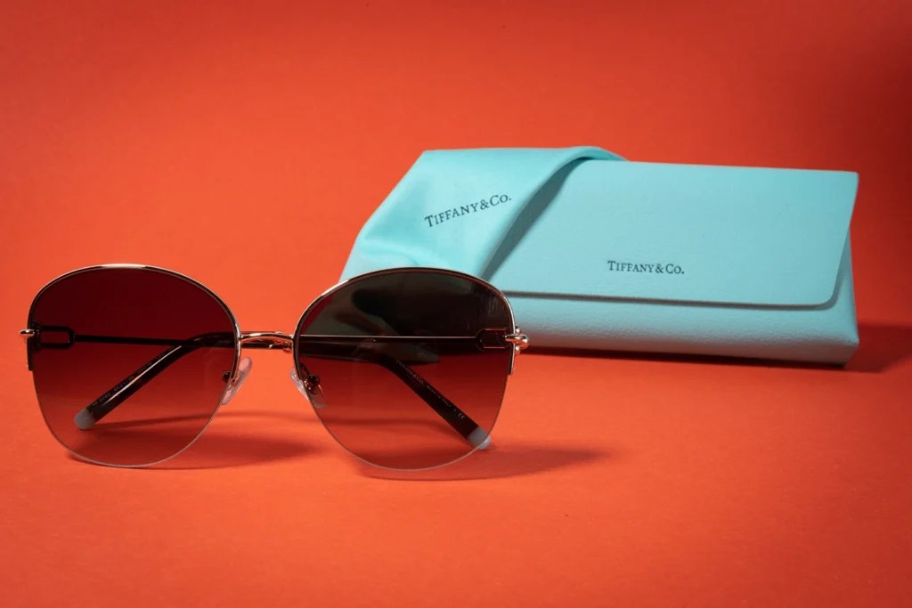 Orangener Hintergrund mit hell-blauer Lederhandtasche von TiffanyCo., darüber hängt ein Brillenputztuch in der selben Farbe mit dem selben Schriftzug. Im rechten Vordergrund befindet sich eine Sonnenbrille