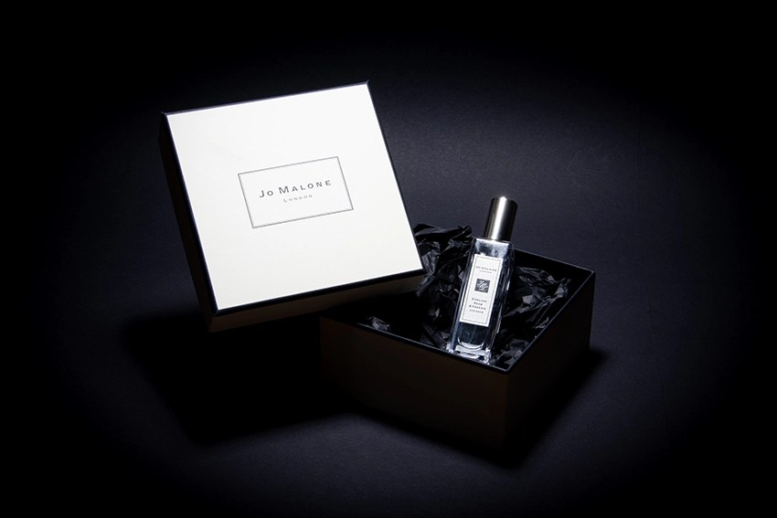 Parfumeflasche von Joe Malone in offener Verpackungsschachtel auf schwarzen Hintergrund. Neben der Flasche befindet sich der Deckel von der Verpackung in der Farbe weiß und mit dem Jo Malone Logo