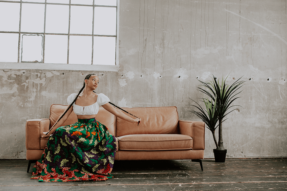 Frau sitzt auf einer braunen Ledercouch und hält ihr langen Zöpfe in den Händen, Sie trägt ein traditionelles afrikanisches Gewand, in einem Industriegebäude