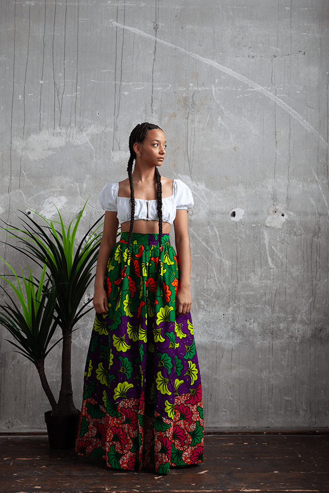 afrikanische Frau mit langen geflochtenen Zöpfen, Sie trägt ein traditionelles afrikanisches Gewand, in einem Industriegebäude. Sie steht vor einer Betonwand neben einer Pflanze und blickt nach rechts