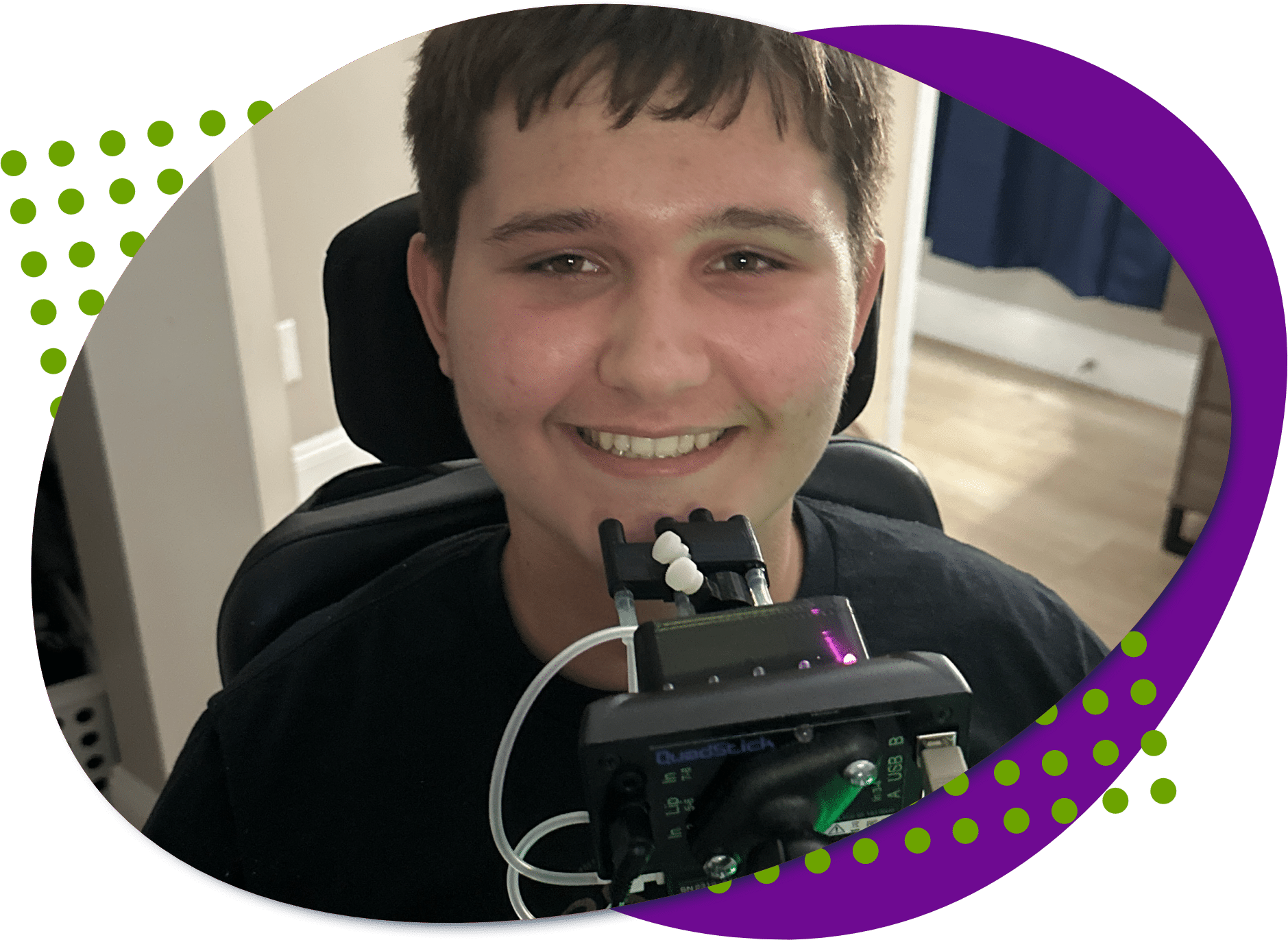 A photo of Carson, he has short dark hair, brown eyes and wearing a black shirt. He is smiling and in front of him is a QuadStick adaptive controller.