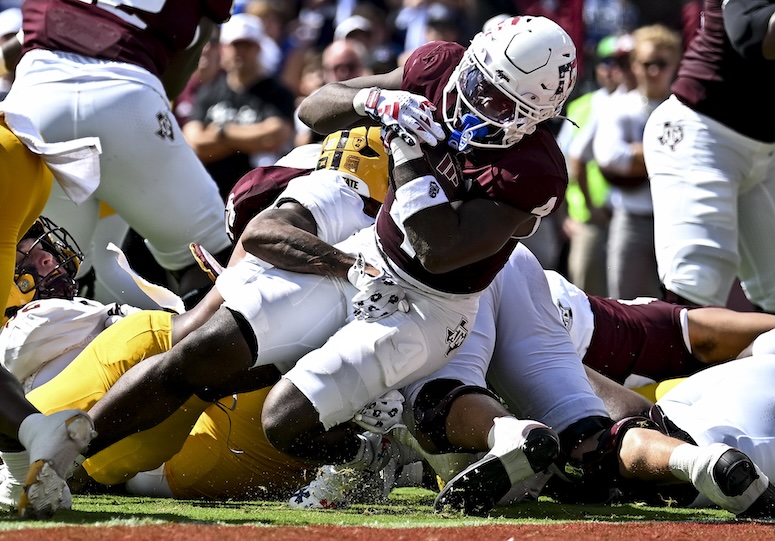 A football player in a maroon jersey is being tackled during a game, with players from both teams on the ground around him.