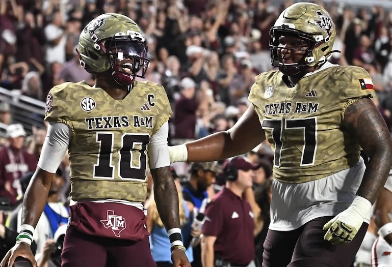 Two Texas A&M football players celebrate on the field, wearing camouflage jerseys and helmets. One player, number 10, appears to be the quarterback, while number 77 offers support. The background features a cheering crowd.