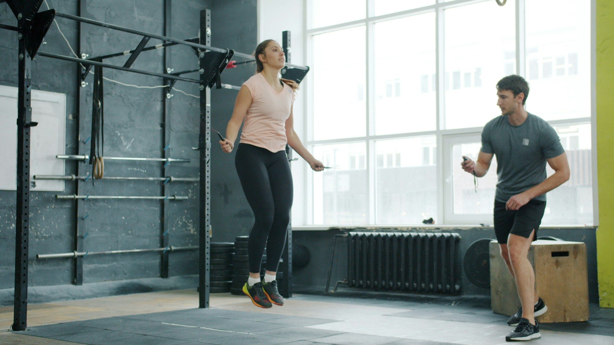 A woman jumps rope in a bright gym while a trainer stands nearby watching
