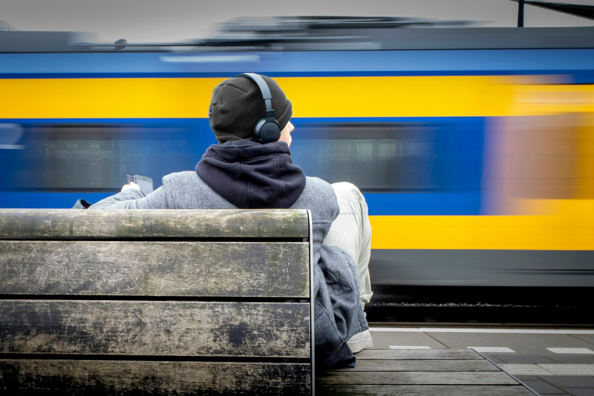 Person wearing over-ear headphones on a station bench as a yellow and blue train blurs past
