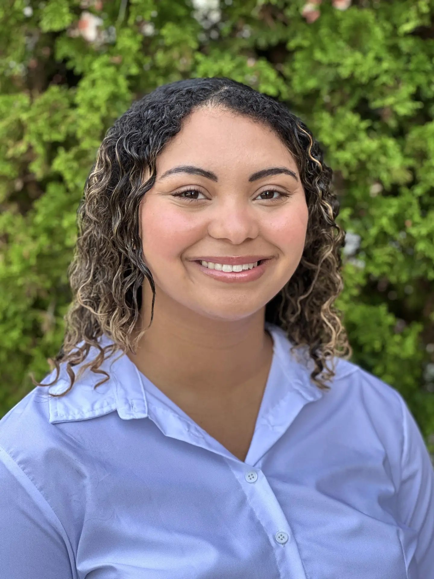 A woman with dark curly hair is shown wearing a light blue shirt and standing against a leafy background