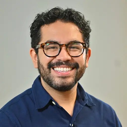 A bearded man with short curly hair and glasses is shown in a dark blue shirt against a gray background