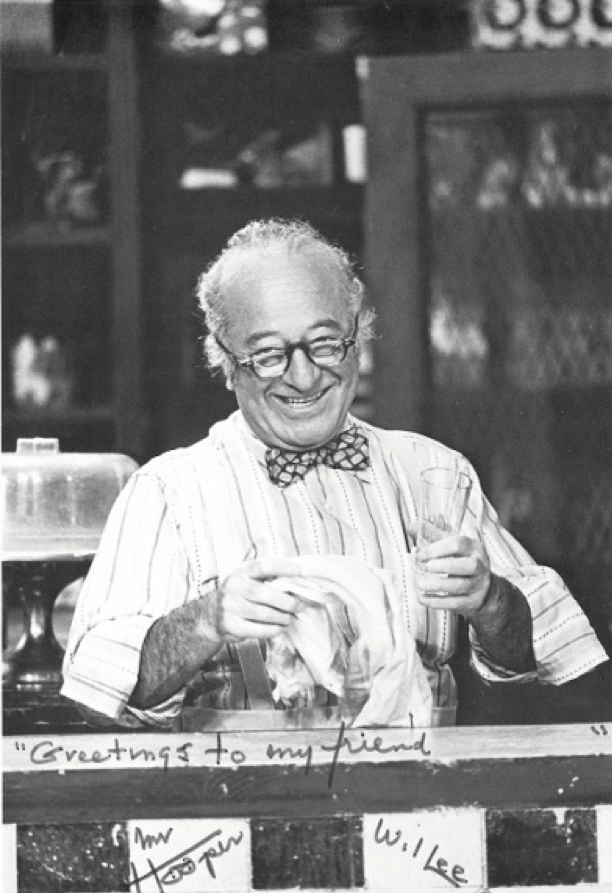 man standing behind table/bar inside and holding a glass and towel.