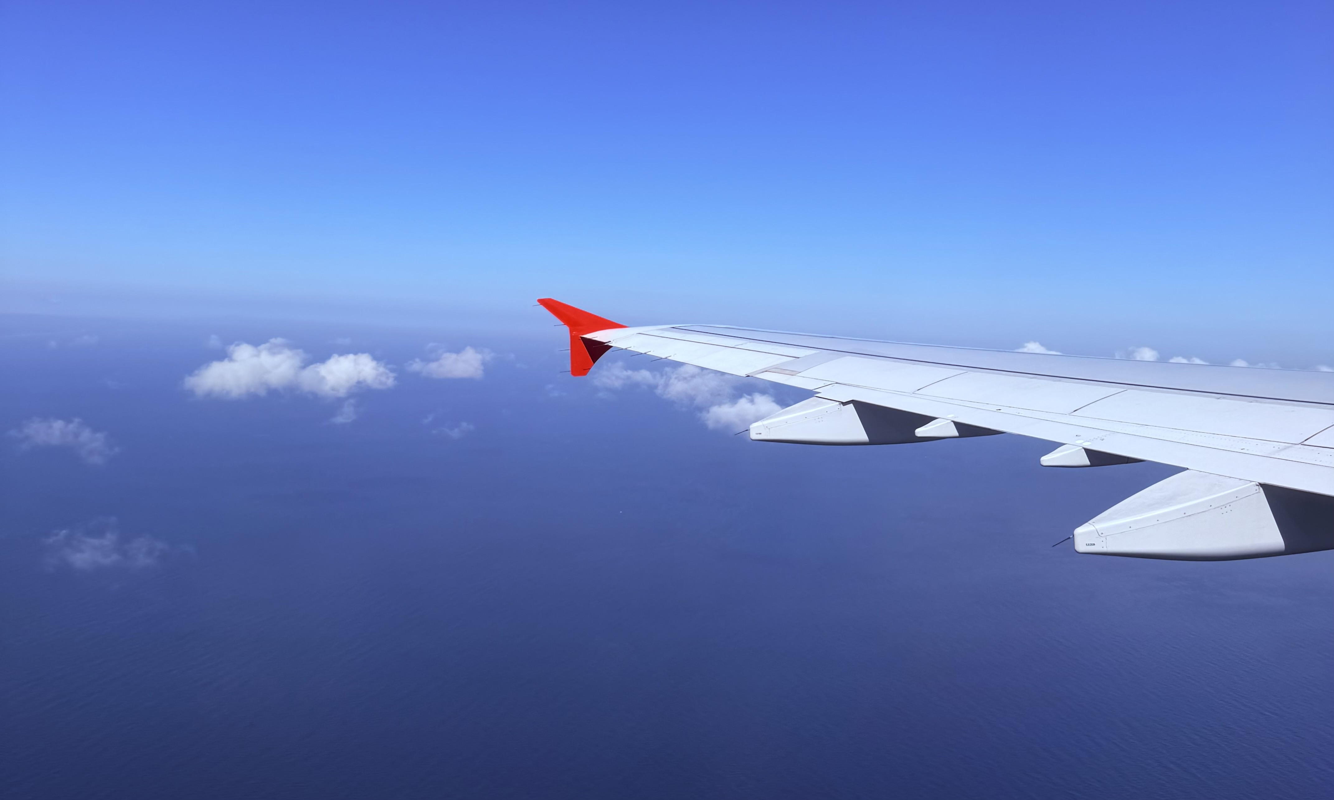 In this image, a red-tipped white plane wing is seen under a deep blue sky, gliding over a vast sea with light white clouds above it.