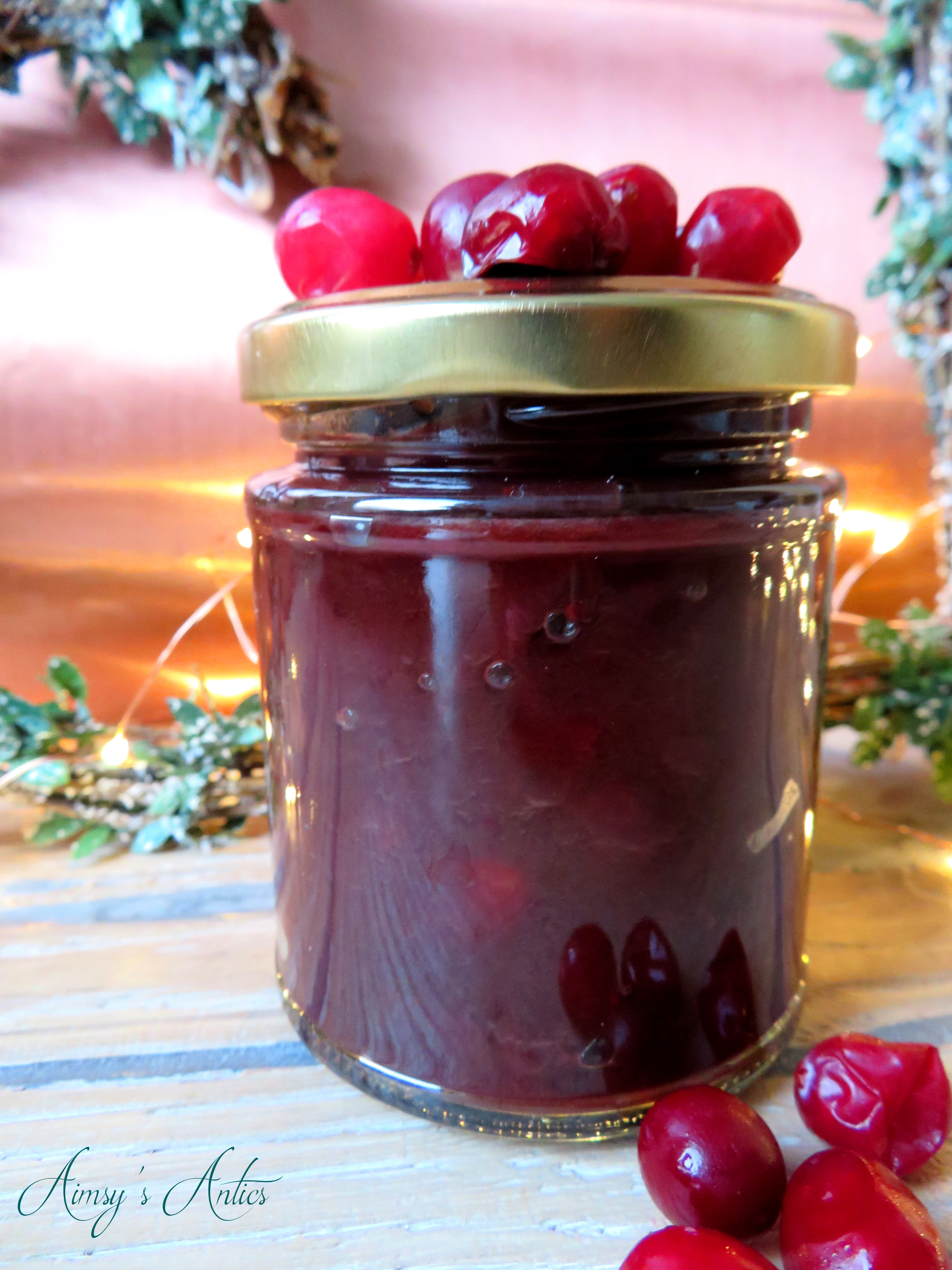 Image of a jar of cranberry and red onion relish, with cranberries on the lid and scattered in front. Christmas heart wreath and fairy lights behind the jar.