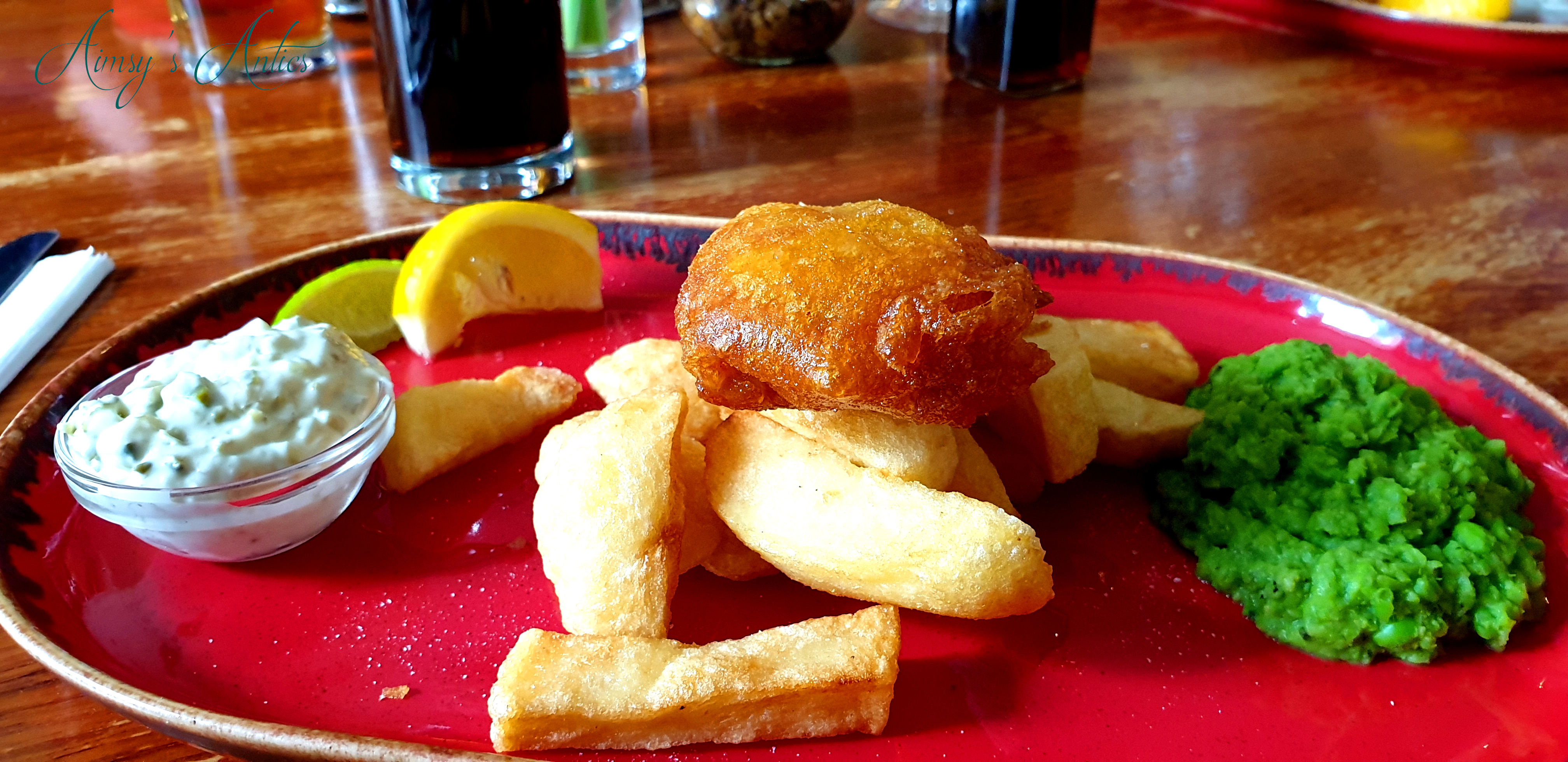 Fish and chips with slightly mashed peas and tartar sauce in a small dish on a red plate.