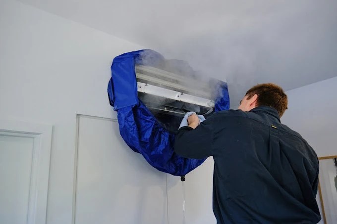 A technician cleaning an indoor air conditioning unit with a cloth, with smoke or vapour surrounding it.