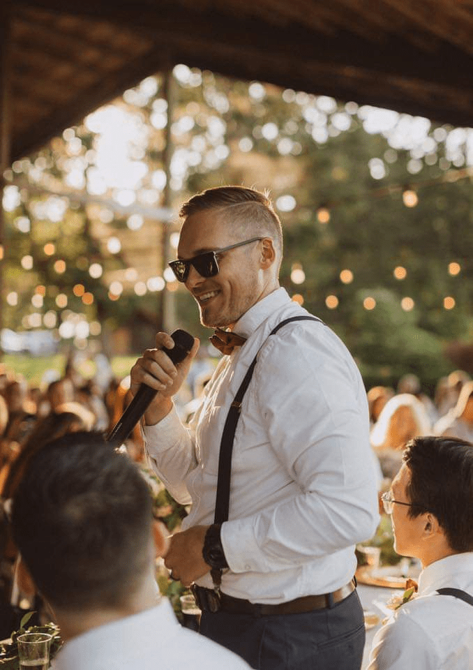 Candid photo of a man speaking into a microphone at an outdoor event, wearing sunglasses, a bow tie, and suspenders