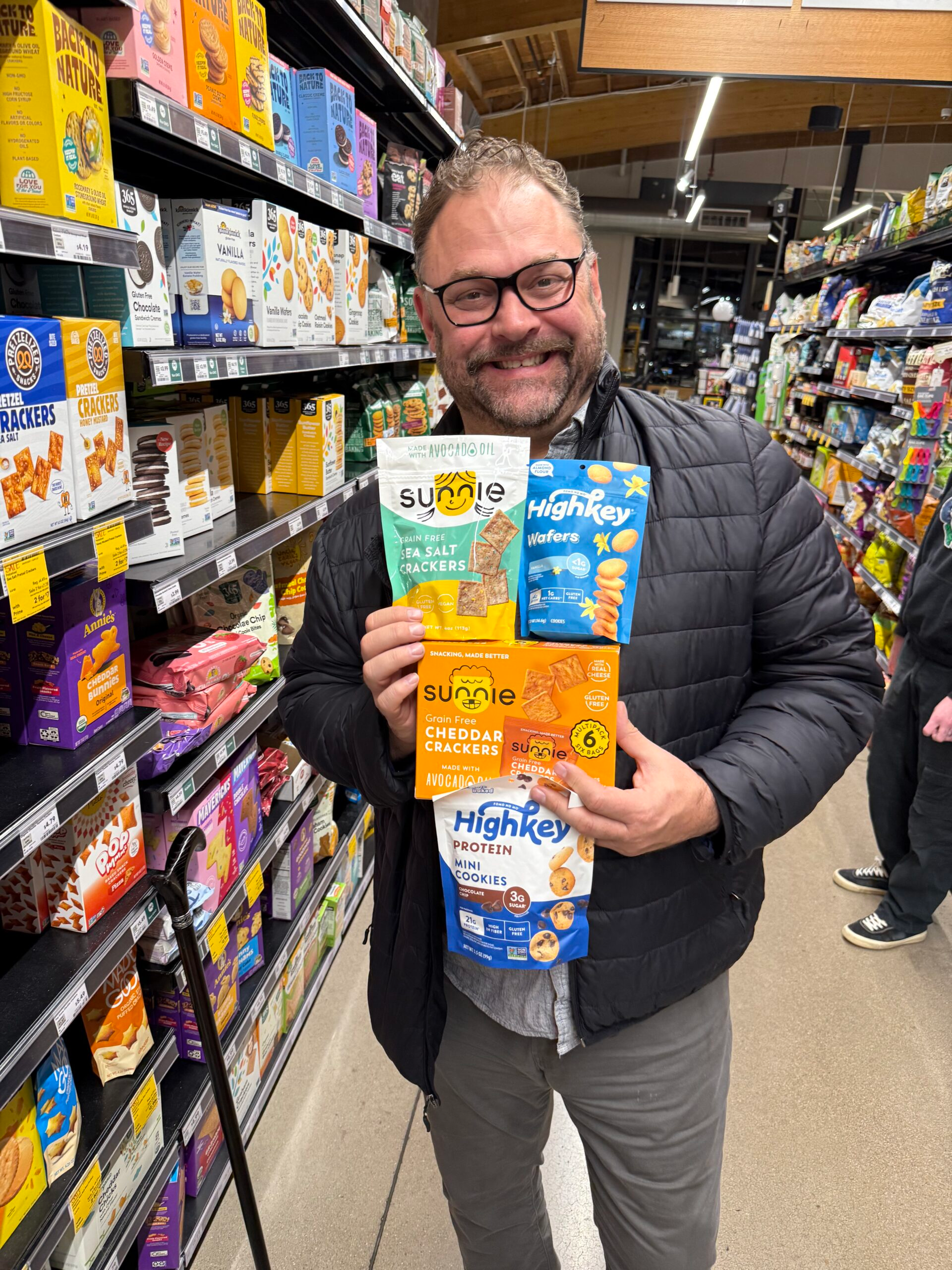 Gabriel Claycamp smiling in a grocery store aisle while holding several packages of grain-free crackers and protein snacks.