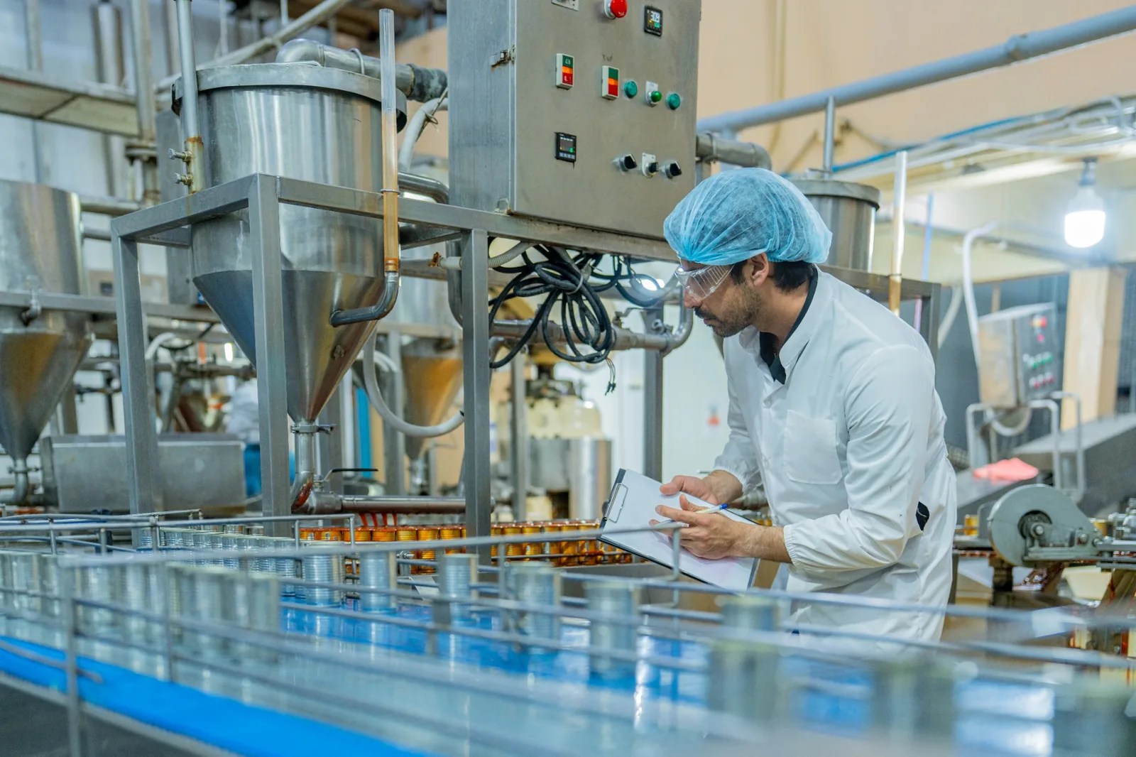 Worker in a hairnet inspects food processing equipment while holding a clipboard.