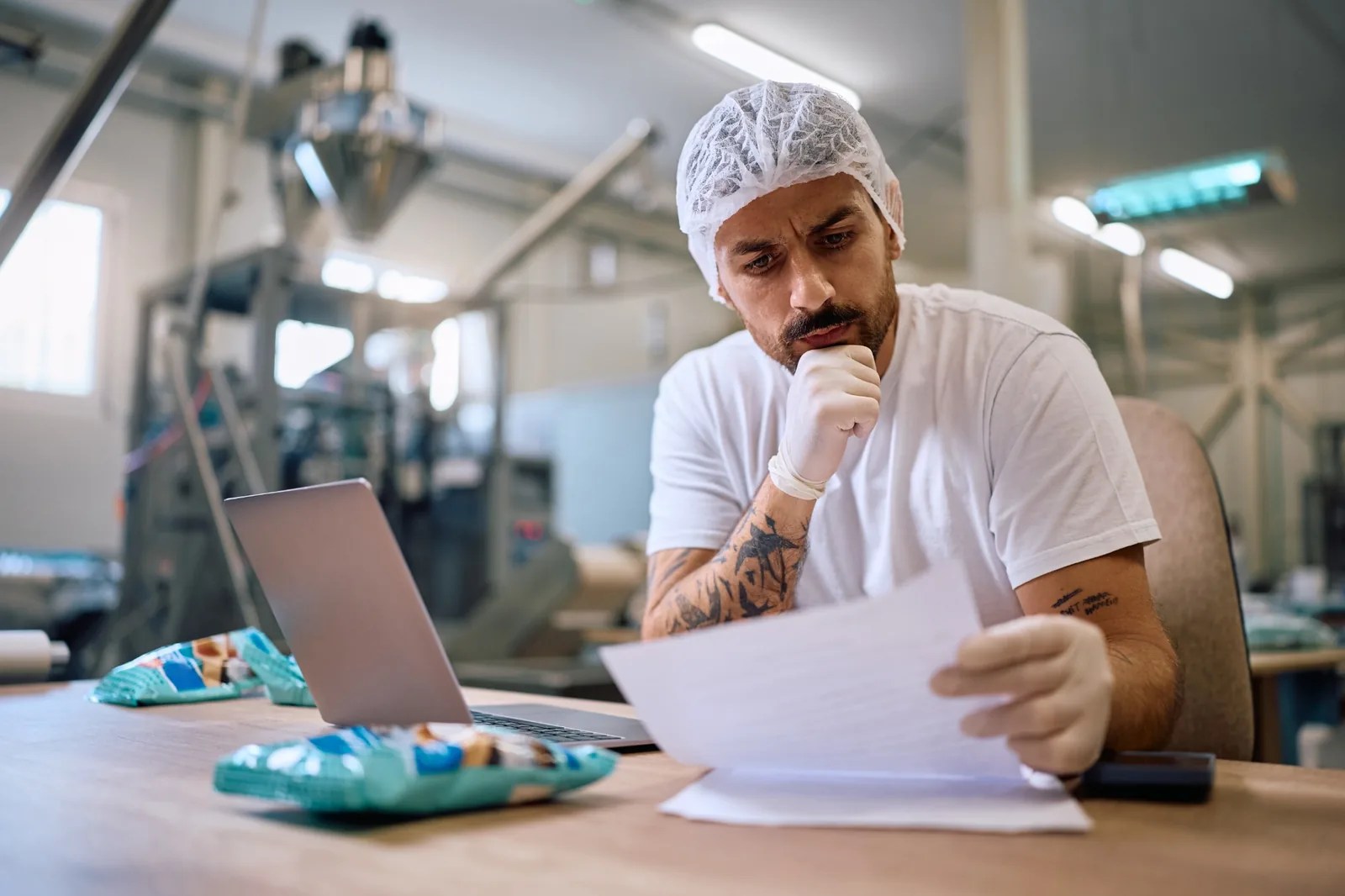 Worker in a hairnet reviews paperwork beside a laptop in a food processing facility.