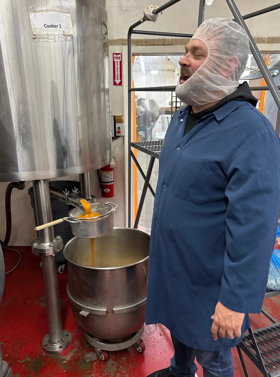 Team member in a blue coat and protective hair covering beside a stainless steel kettle dispensing orange liquid through a strainer.