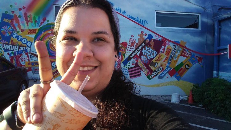 About Alethea Rocks - image /selfie of Alethea holding a cup while giving a peace sign in front of candy wall that can be found in Wilton Manors behind To the Moon Marketplace back entrance