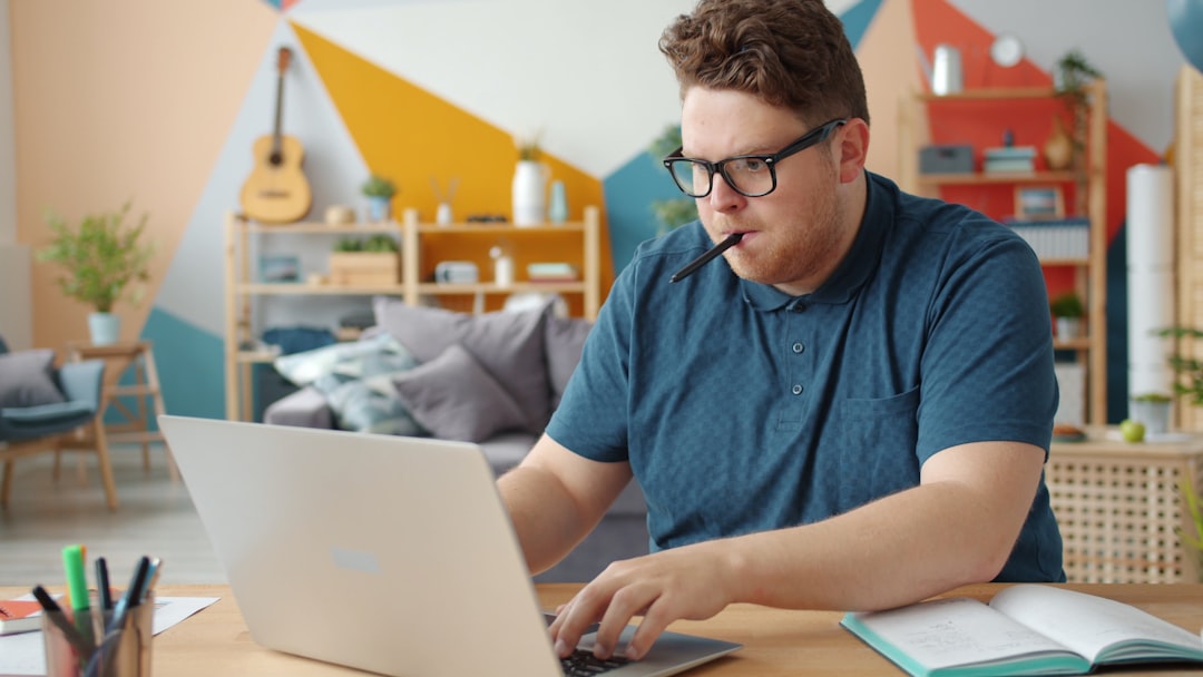 A male student holding a pen in his mouth sits at a table in his home, looking at a computer with a notebook open next to him.