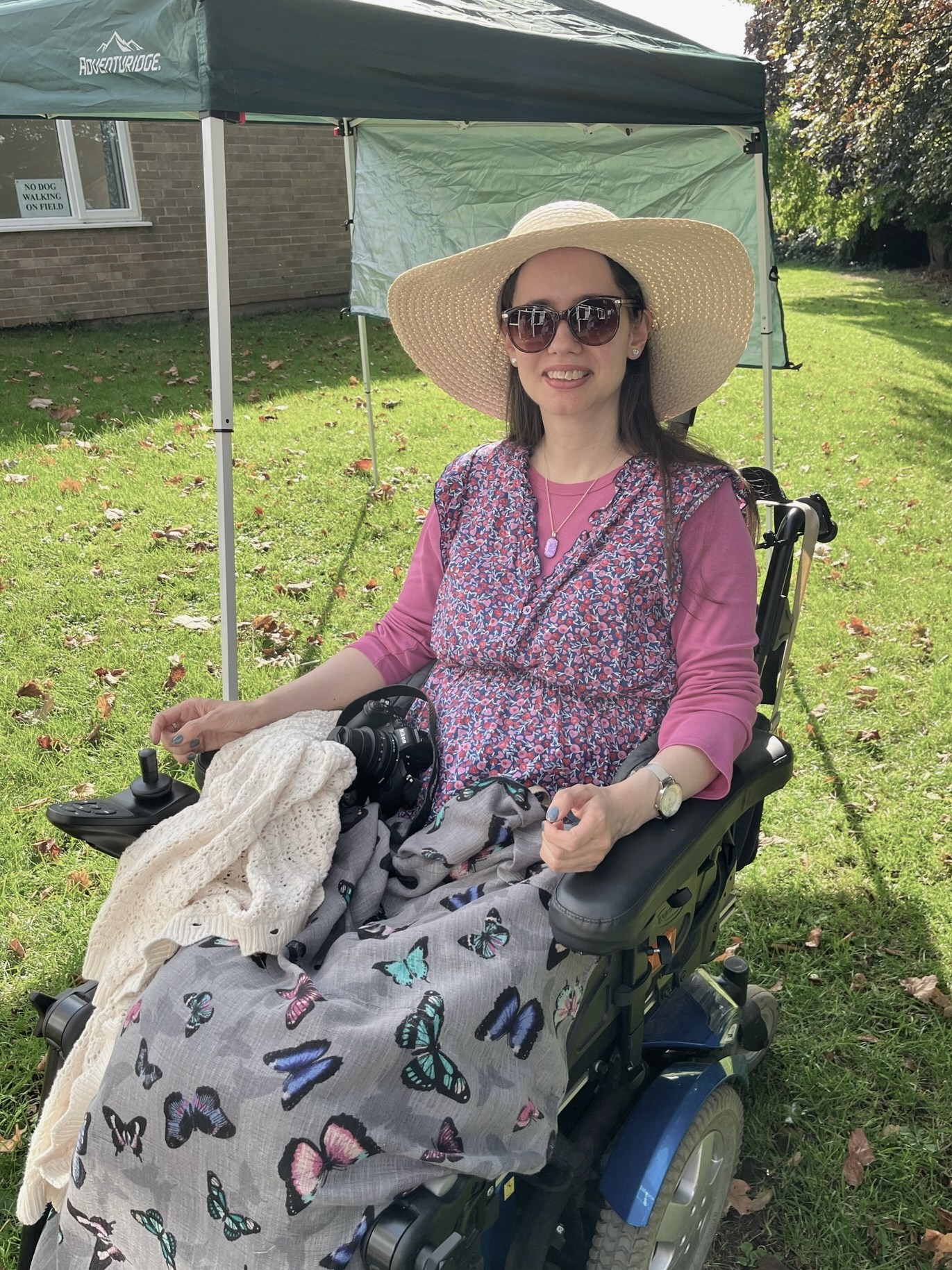 Portrait of the author, wearing a pink shirt and floral vest and sitting outside in a wheelchair