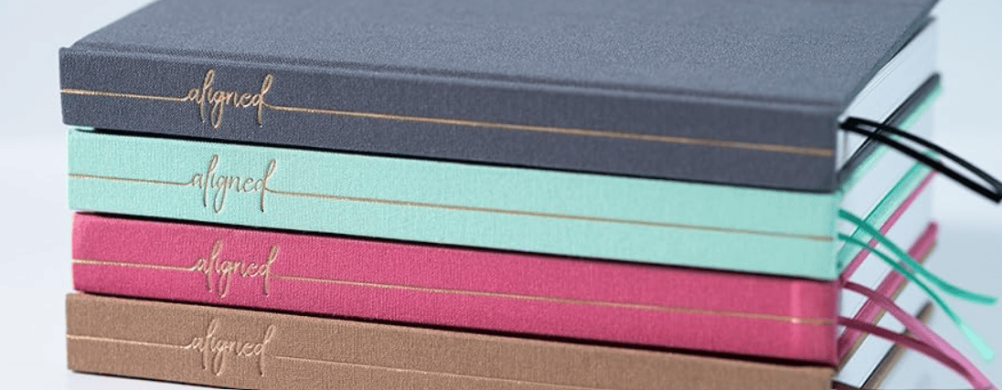 A stack of notebooks in various colors including gray, mint green, pink, and brown, with the word 'aligned' embossed in gold on each cover.