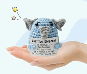 A small, crocheted blue elephant held in a hand, with a tag that reads 'Positive Elephant' along with an encouraging message.