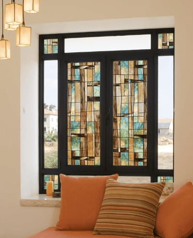 Stylish window with stained glass featuring geometric patterns in blue, amber, and brown, set within a black frame, overlooking an outdoor landscape. A cozy seating area with an orange cushion and striped pillow is visible in the foreground.