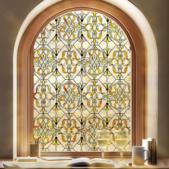 Stained glass window featuring intricate floral patterns in warm hues of yellow, orange, and black, framed by a wooden arch, with a table showcasing drinks and a book in the foreground.