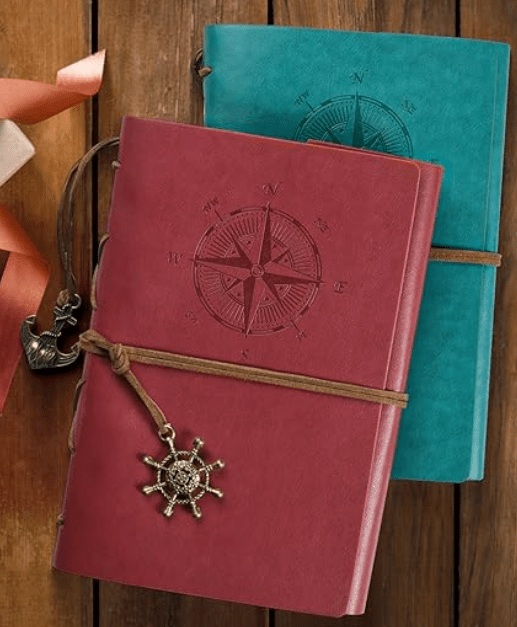 Two leather-bound journals with embossed compass designs, one in red and the other in teal, tied with brown string and adorned with a decorative ship's wheel charm, placed on a wooden surface.