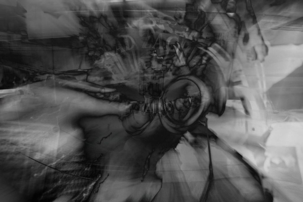 Black-and-white layered photo-montage with large goggle-like lenses at the centre, surrounded by blurred architectural and mechanical forms streaking outward in radial motion.