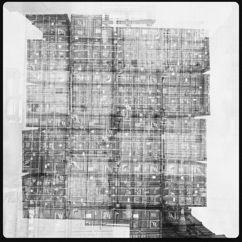 Black-and-white layered photograph of stacked shipping containers forming a broad, subtly head-like mass, covered with repeated corrugation, numbers, labels and shipping symbols.
