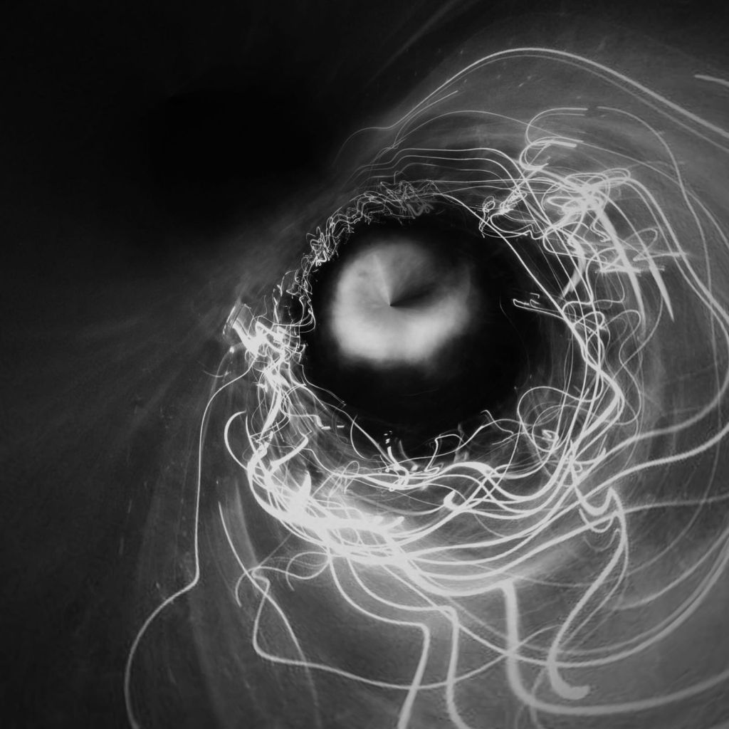 Square black-and-white abstract photograph with a dark circular centre surrounded by tangled, orbiting white light trails. A softly illuminated form sits within the centre, while the left side remains largely black.