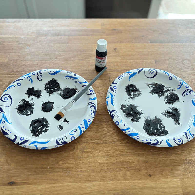 Two paper plates sitting on a table with the letters "D", "A", "I", "L", and "Y" on one plate and the letters  "R", "H", "Y", "T", "H", and "M" on the other plate. All the letters have been painted black.