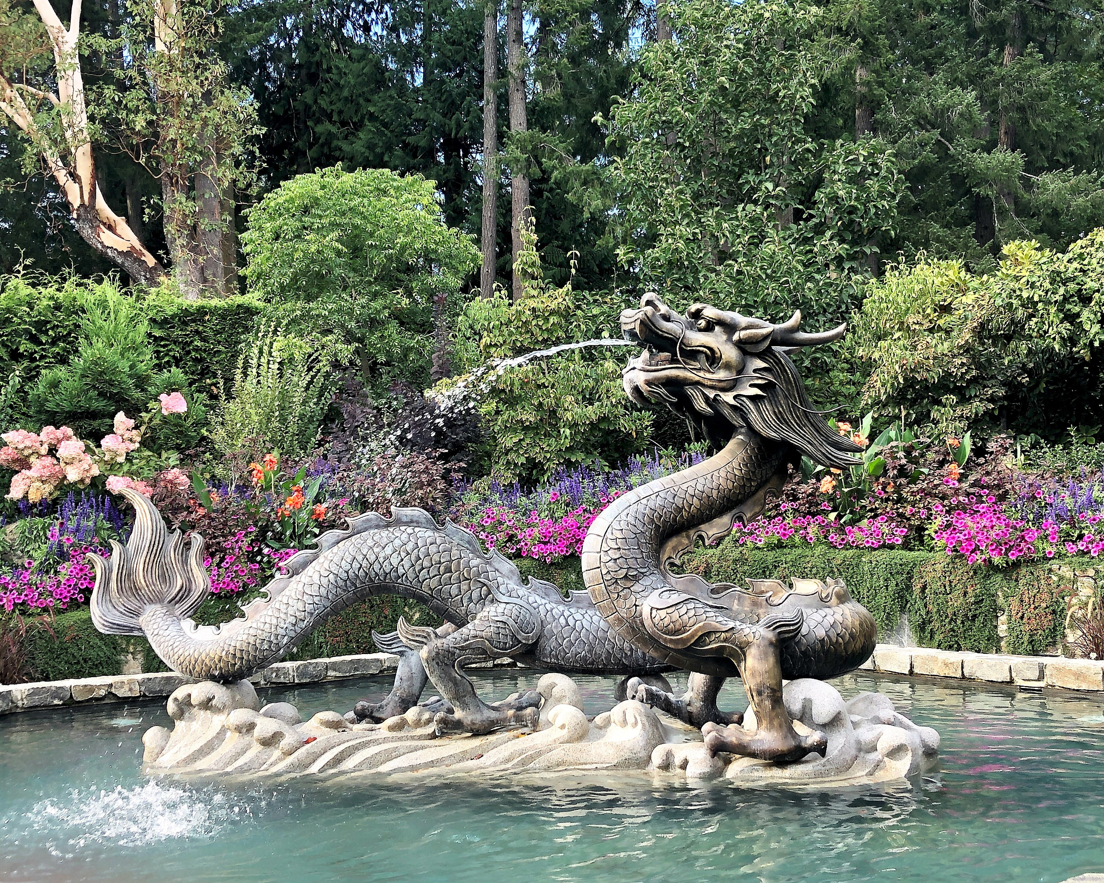 A fountain shaped like a dragon with water spewing from its mouth sits in a pool surrounded by flowers, shrubs and trees.