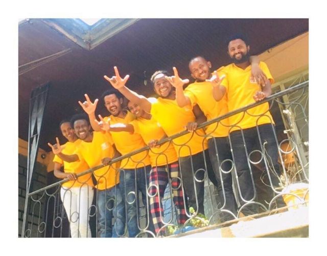 7 people smiling down fro a balcony - all in yellow tshirts
