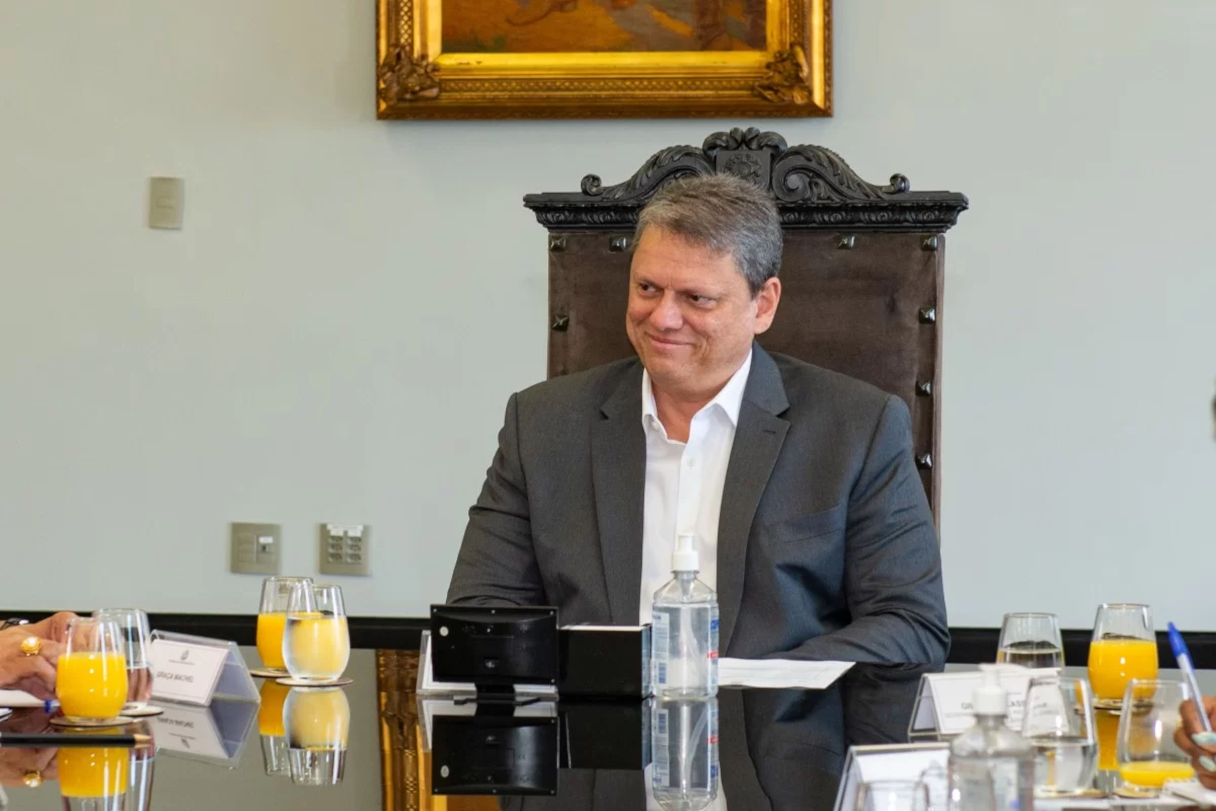 Imagem mostra o governador Tarcísio de Freitas, um homem branco e de terno, sentado à mesa. Há documentos e copos com suco na mesa.