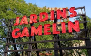 Bright red sign reading “Jardins Gamelin” mounted on a dark wooden frame, with leafy green trees and clear blue sky in the background. This popular public space near downtown supports the area’s appeal as one of the best places to stay in Montreal for older travellers.
