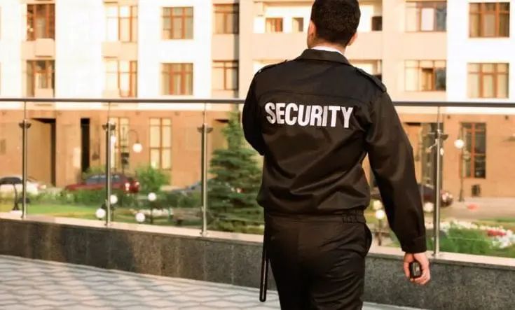 Security guard in black uniform with 'SECURITY' written on the back, patrolling outside a building.