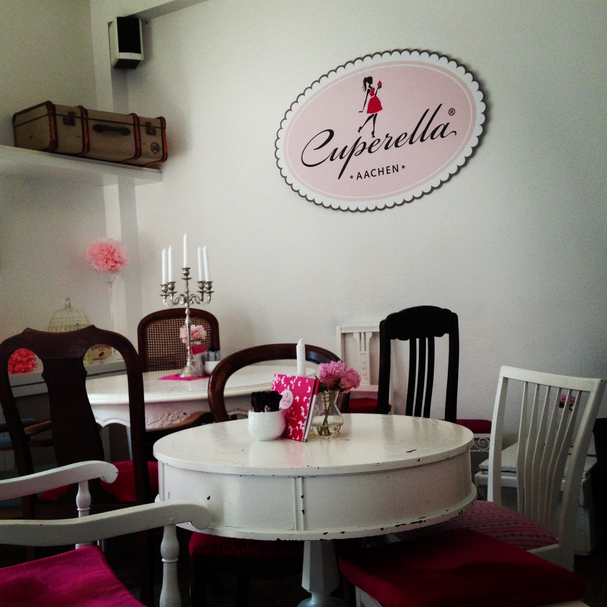 Das gemütliche Café-Interieur verfügt über einen runden weißen Tisch, der von ungleichen Stühlen umgeben ist. Rosa Kissen und rosa Blumendekor betonen den Raum. Auf einem Regal darüber steht ein Vintage-Koffer. An der Wand hängt ein Schild mit der Aufschrift „Cuperella Aachen“ und die Silhouette einer Frau, die einen Cupcake hält.