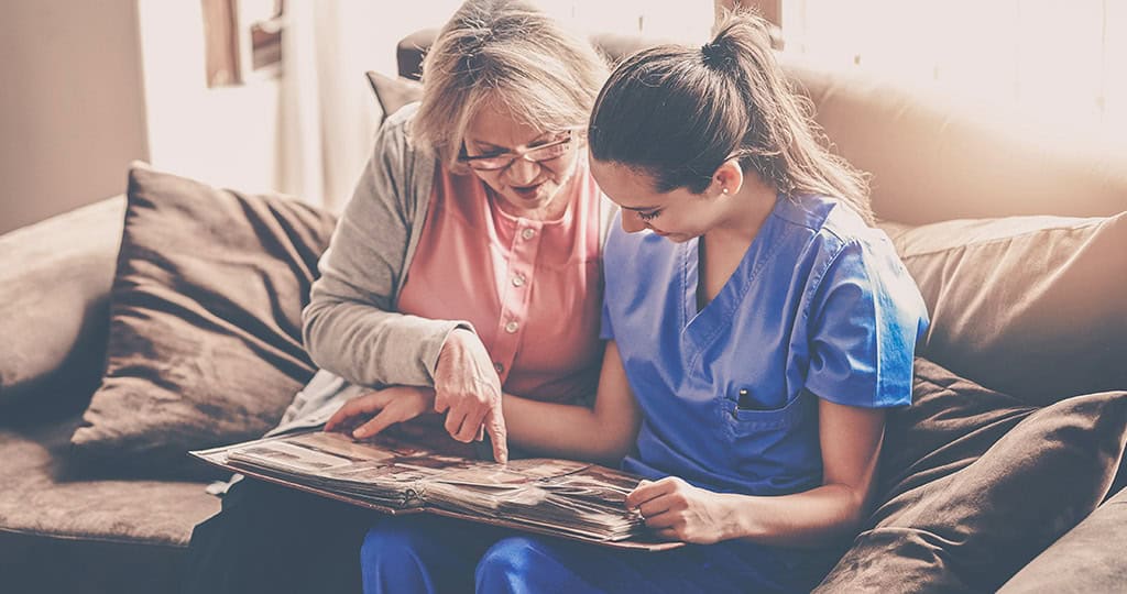 Healthcare worker and an elderly woman smiling while looking at a photo album together on a sofa in elderly home care.