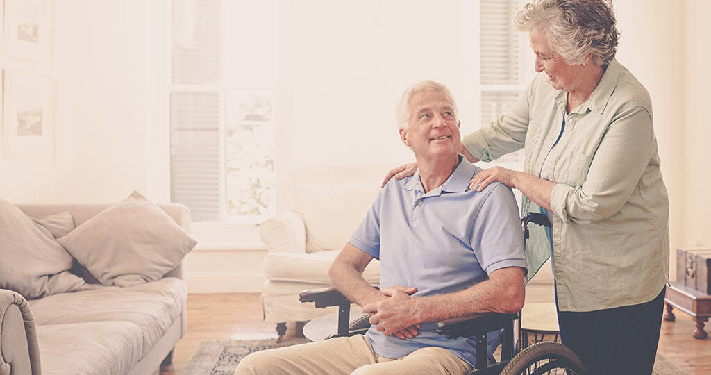 Senior woman standing next to husband in a wheelchair with her hands on his shoulders