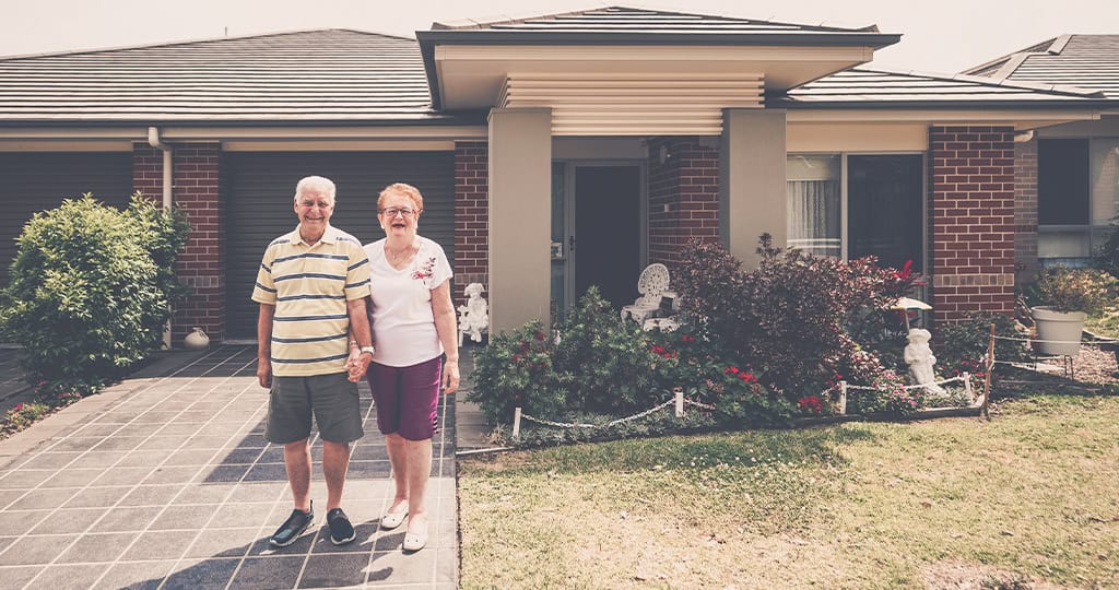 Senior couple in front of their suburban home with a manicured garden, emphasizing home care.