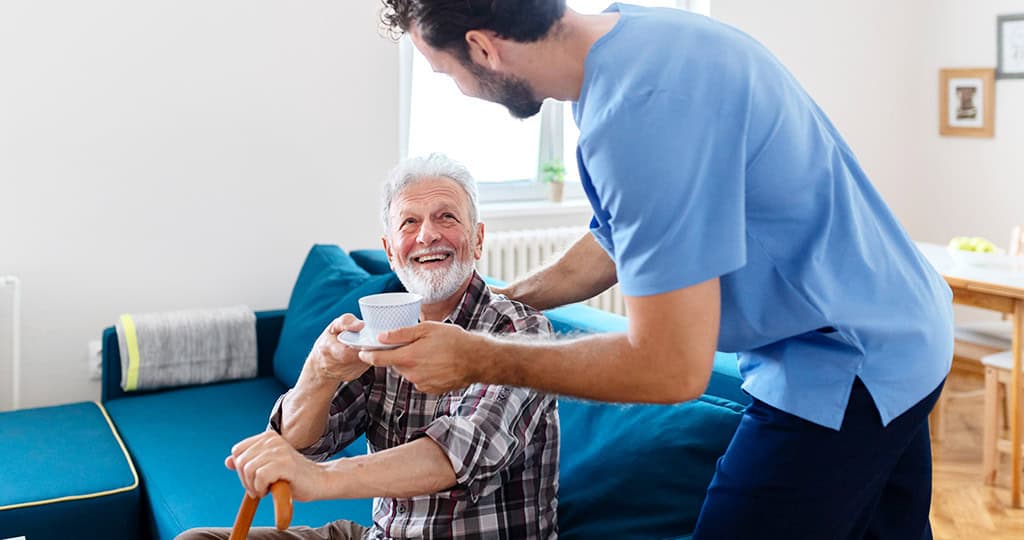 Caregiver handing tea to an elderly man sitting on a couch with a cane